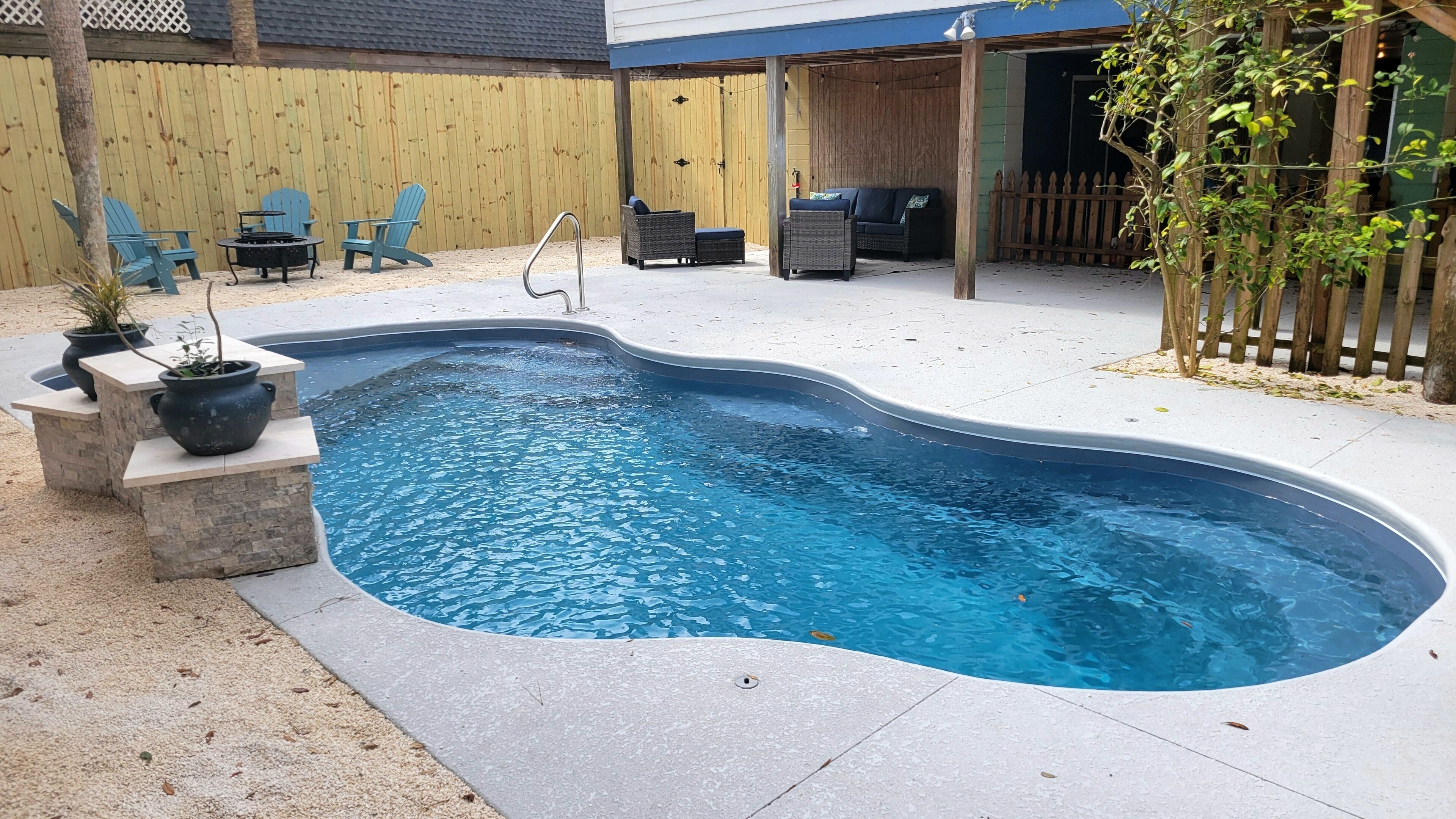 A swimming pool with a stone deck, surrounded by a wooden fence, and outdoor furniture.