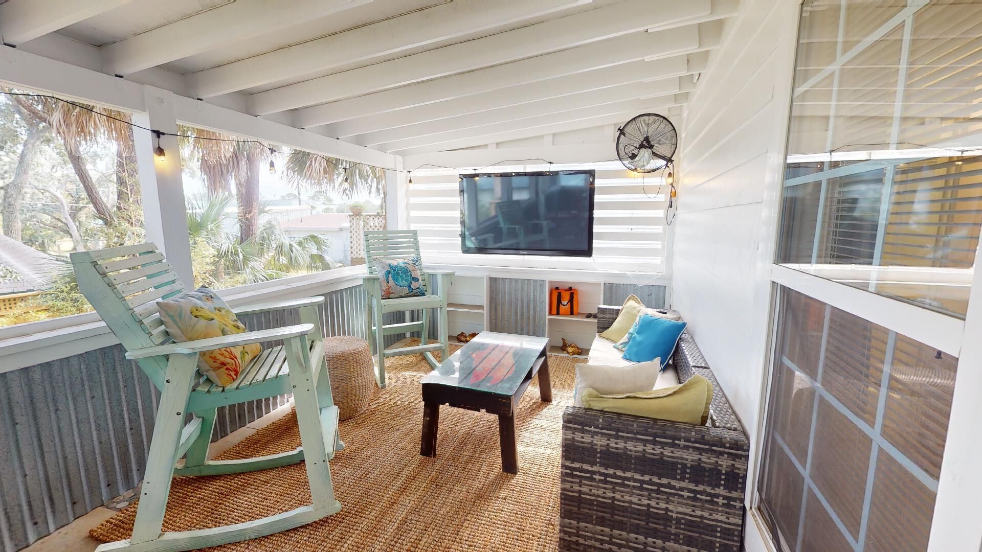 Un espacio de estar al aire libre cubierto, con televisor, mobiliario de mimbre y vistas al exterior.