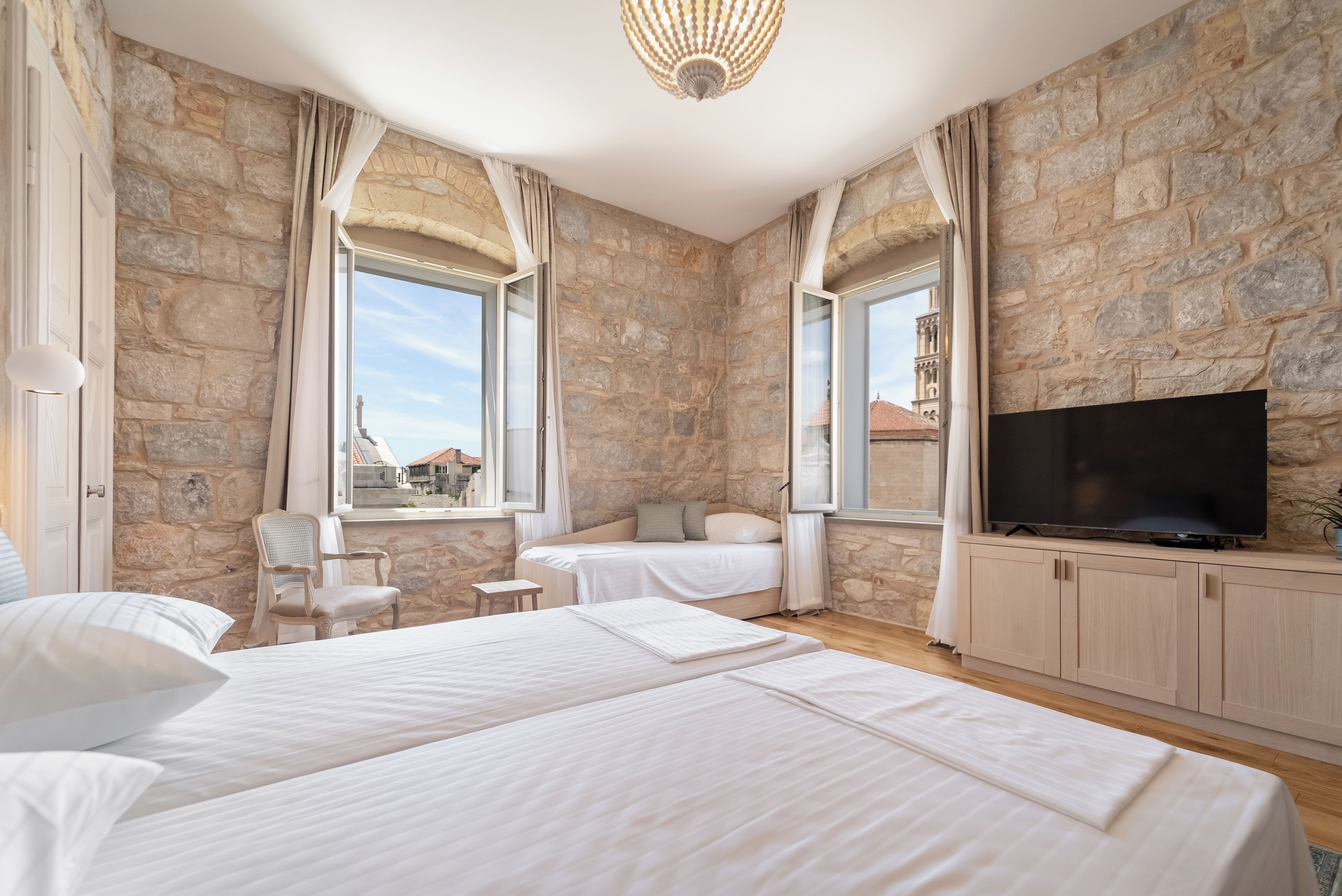 A hotel room with stone walls, a large TV, two beds, a chair, and a window with a view.