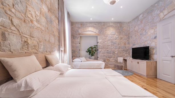 Un dormitorio con paredes de piedra, una cama con ropa blanca, un televisor sobre un mueble de madera y una planta en la esquina.