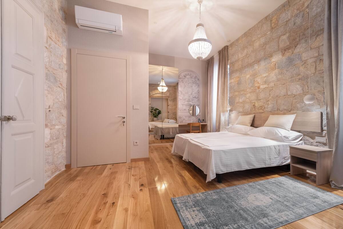 Un dormitorio con pared de piedra, suelo de madera, una cama con ropa blanca, una mesita de noche, una araña y vista a otra habitación.