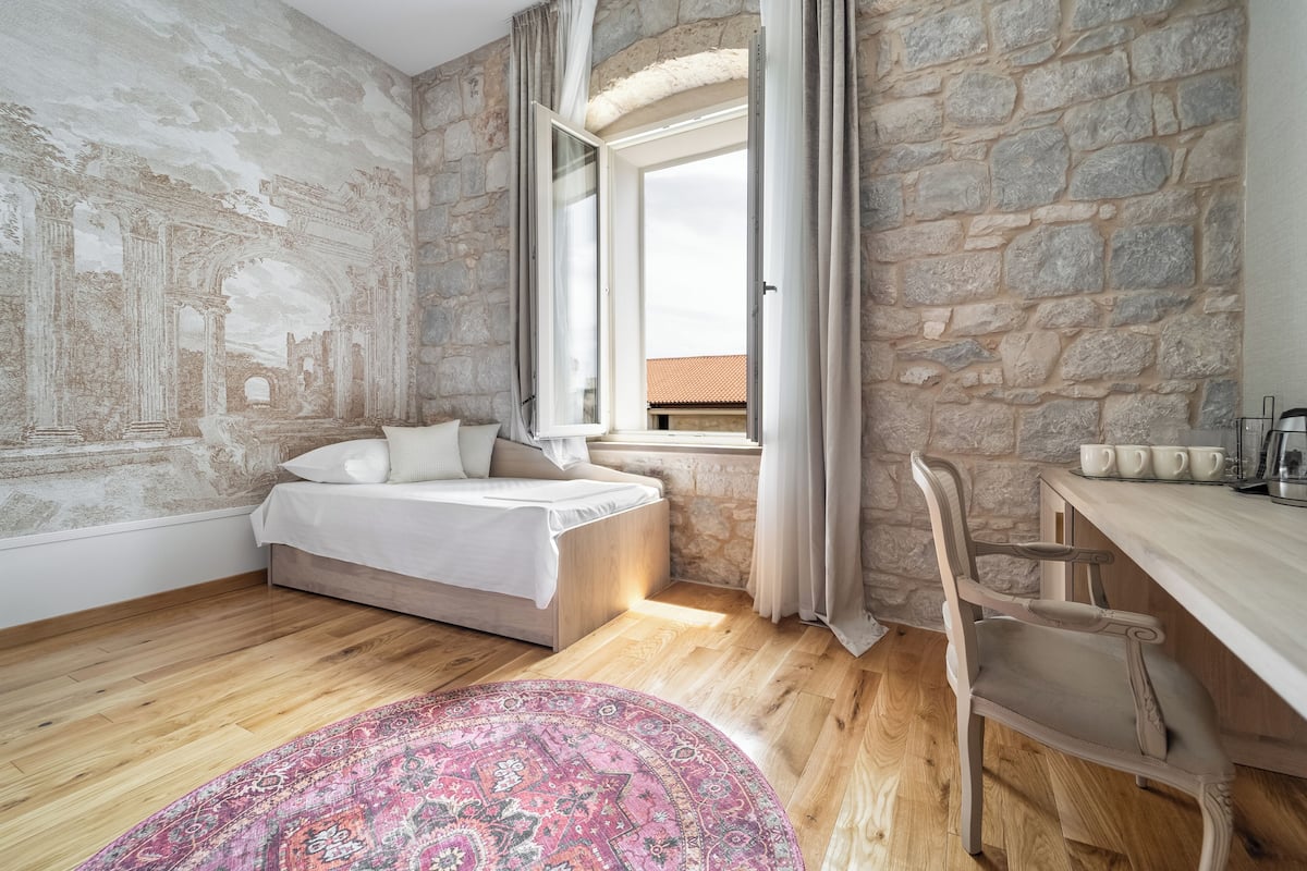Un dormitorio con pared de piedra, una cama, un ventanal con cortinas, suelo de madera y alfombra estampada.
