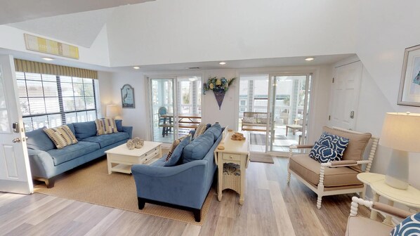 Una sala de estar amplia con un sofá azul, un sofá color beige y una mesa de centro. Se ve un comedor con una mesa y sillas a través de ventanas grandes.