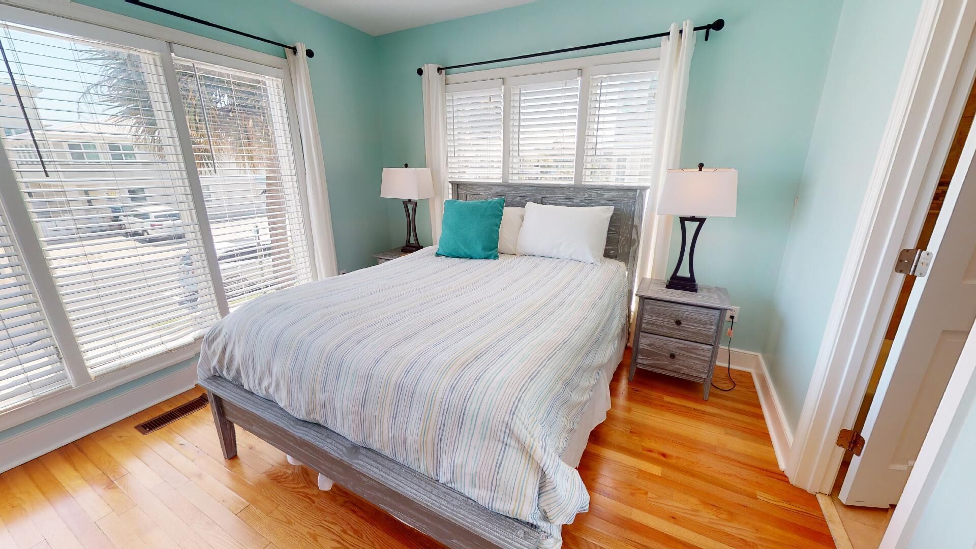 A bedroom with a bed, two bedside tables, a window with blinds, and a door.