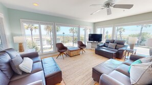 A spacious living room with a large TV, comfortable seating, and a view of the beach.