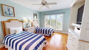 A bedroom with two beds, a ceiling fan, a large window with blinds, and a TV on a dresser.