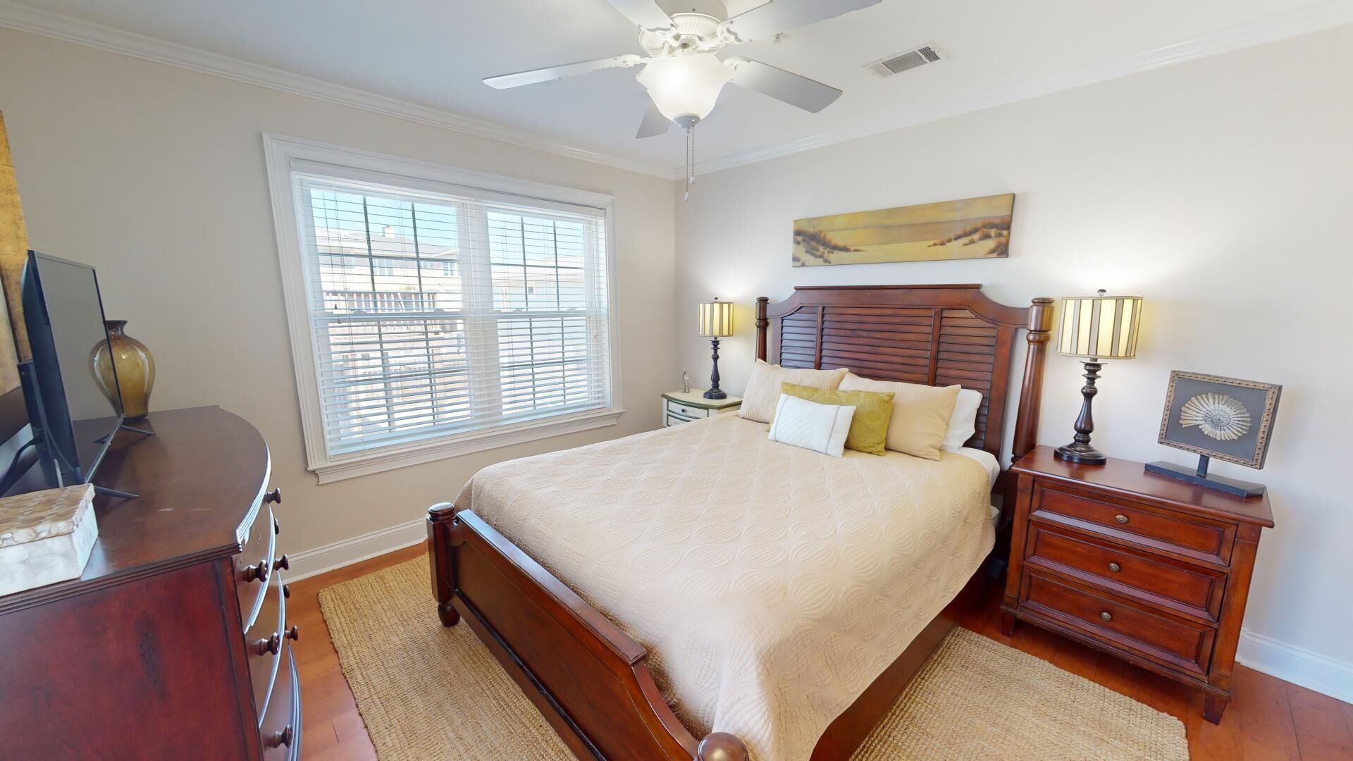 A bedroom with a large bed, a ceiling fan, a window with blinds, a nightstand with a lamp, and a dresser.