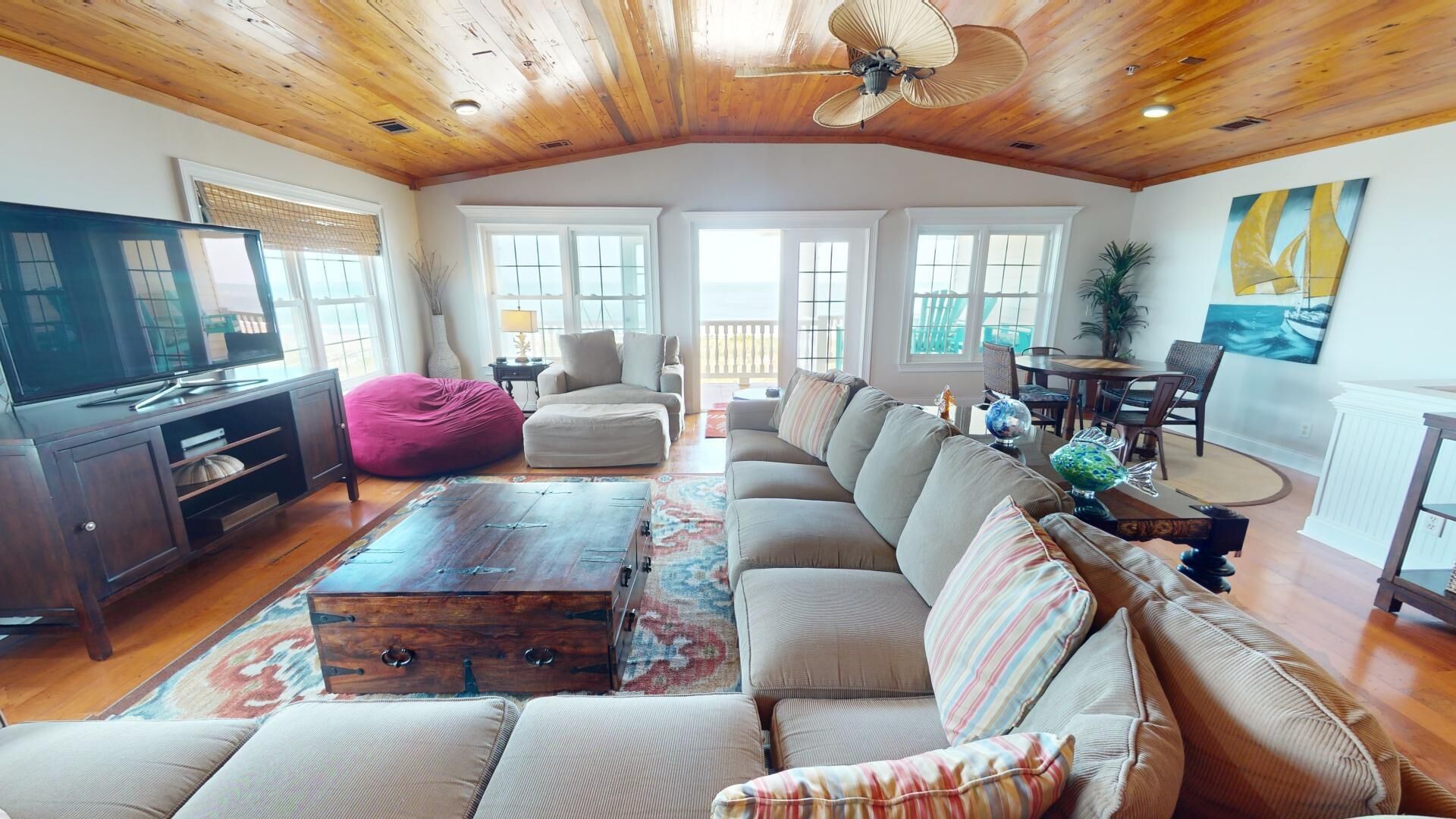 A spacious living room with a wooden ceiling, a large sofa, a bean bag, a TV, and a dining area with a table and chairs.