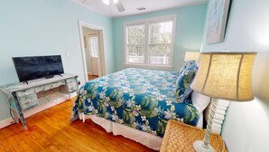 Une chambre à coucher avec un lit, un téléviseur posé sur une commode et une fenêtre avec des stores.