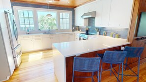 A modern kitchen with white cabinets, a large island with bar stools, and a view of greenery outside. - 3 10th Place by Tybee Vacation Rentals (Tybee Island)