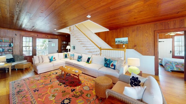 Un salon spacieux avec un grand canapé d’angle, une table basse et un tapis à motifs. Un escalier doté d’une rampe blanche mène à un autre niveau.