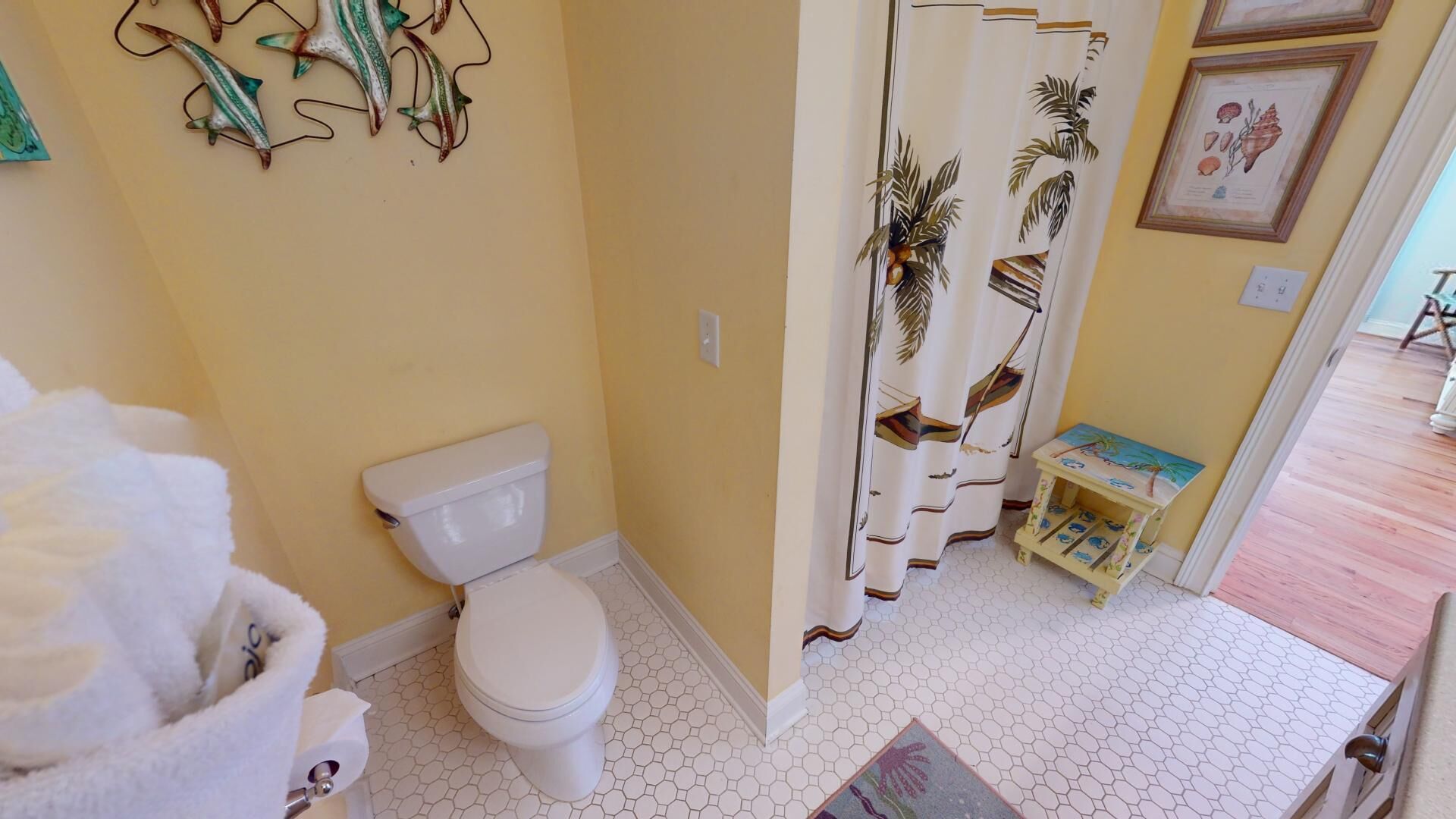 A bathroom with a toilet, a shower curtain with a tropical design, and a small table with a colorful top.