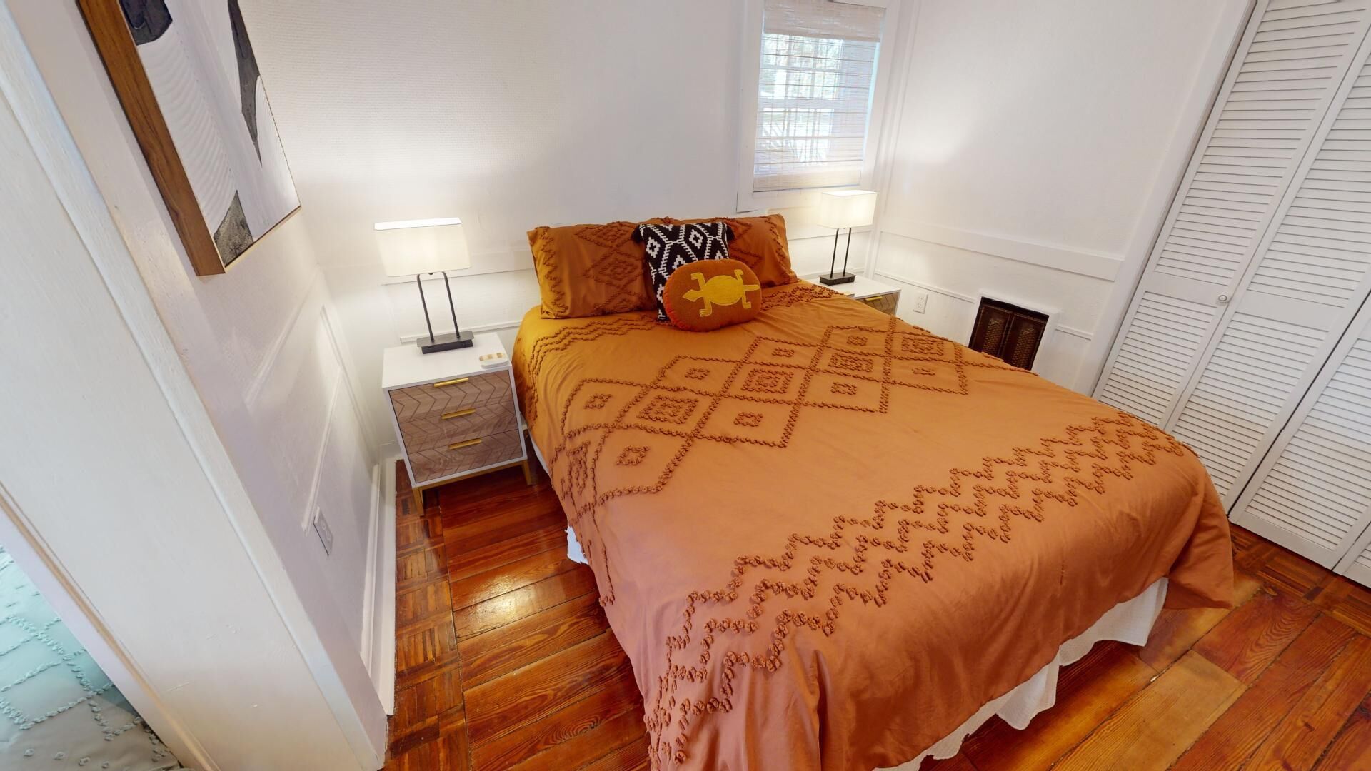 Un dormitorio con piso de madera, una cama con colcha estampada, dos lámparas de noche, una mesita de noche y una ventana con persianas.