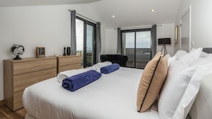Habitació d'hotel moderna amb un llit gran, un taulell de fusta, un escriptori amb una làmpada i vista al mar a través d'una porta corredissa.