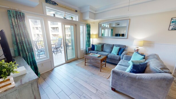 Un vaste salon avec un canapé gris, une table basse et un téléviseur. De grandes fenêtres sont ornées de rideaux, un balcon est visible à travers les portes vitrées, et un miroir est accroché au mur.