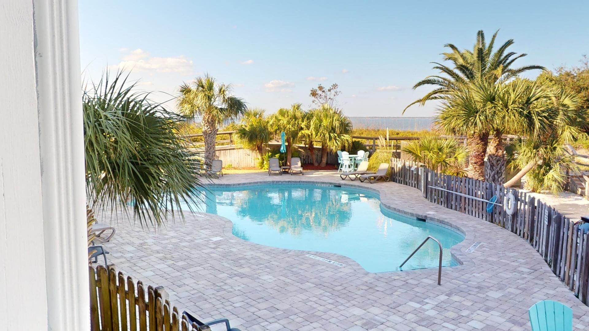 A swimming pool with a curved edge, surrounded by a brick patio and a wooden fence. There are lounge chairs and a small table near the pool.