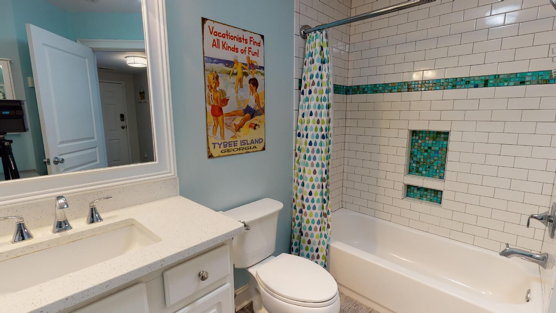 Ein Badezimmer mit einem weißen Waschbecken, einer Toilette und einer Badewanne. An der Wand hängt ein Plakat, das Tybee Island, Georgia, bewirbt.