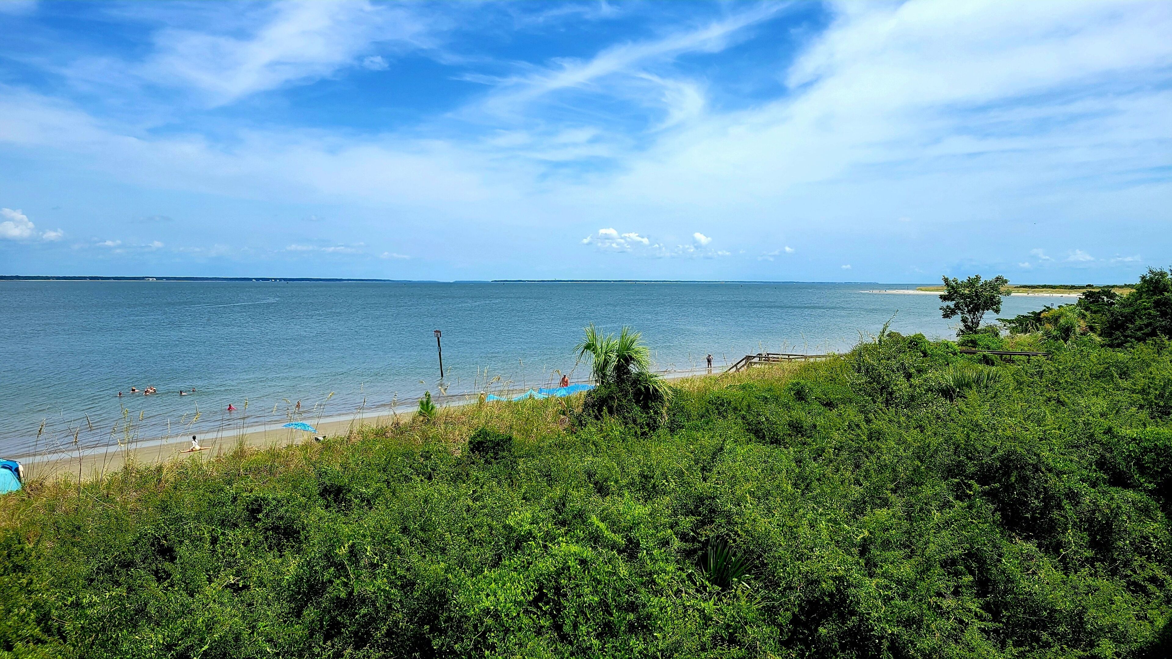 A beach with clear blue water, a sandy shore, and lush greenery in the foreground.