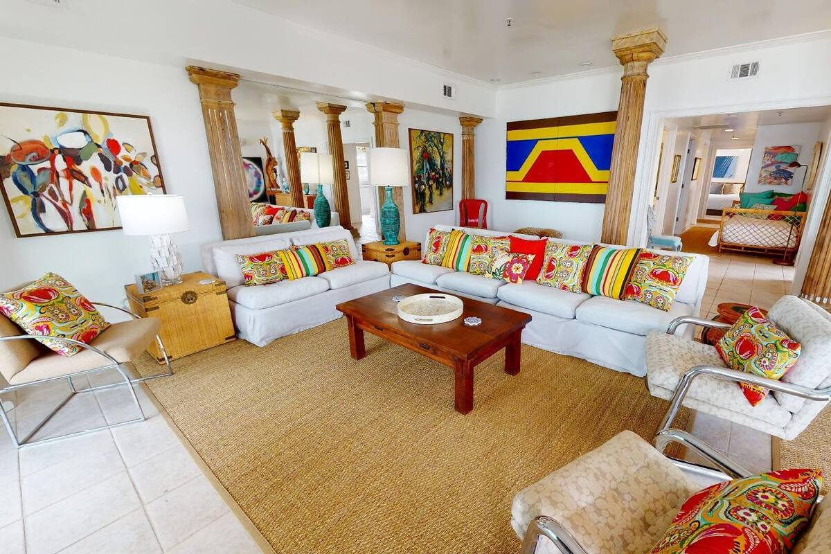 A spacious living room with a white sofa set, a wooden coffee table, and colorful throw pillows. There are large paintings on the walls and a view into another room through an open doorway.