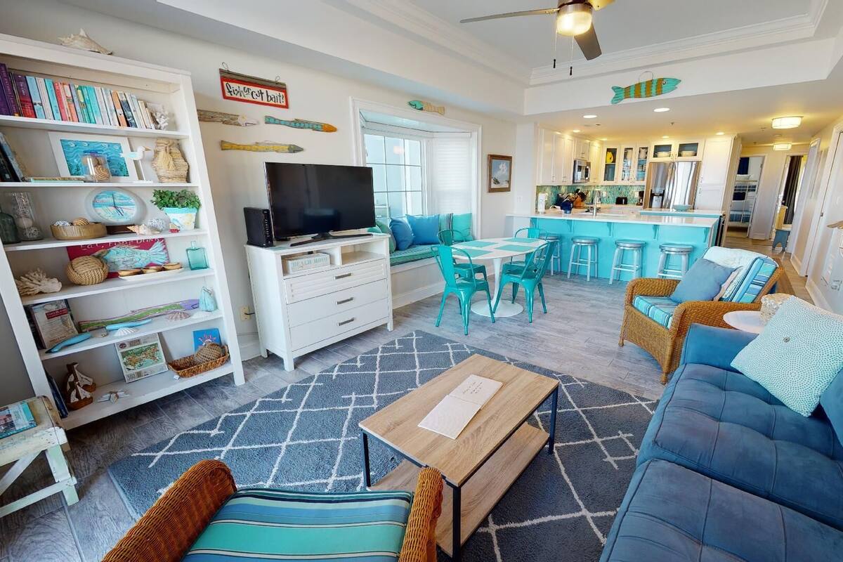 A spacious living room with a sofa, a coffee table, and a TV. There is a kitchen area with a bar and stools.
