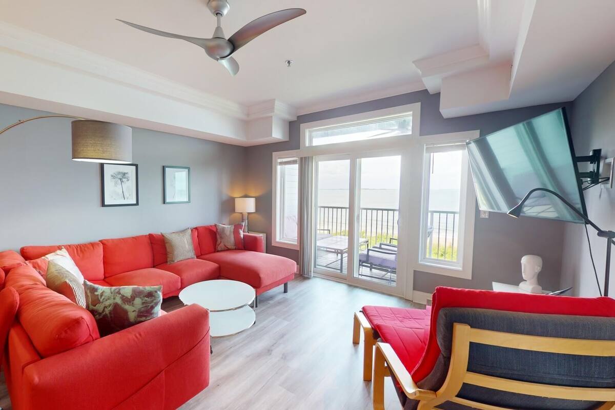 A modern living room with a red sectional sofa, a white coffee table, and a wooden chair. There is a ceiling fan, a floor lamp, and a large window with a view of the outdoors.