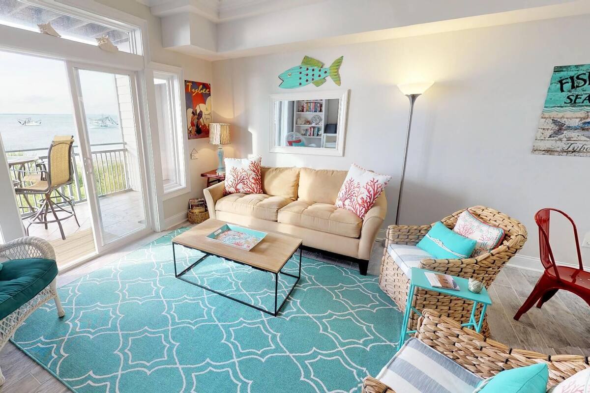 A spacious living area with a large window overlooking the sea, a beige sofa, wicker chairs, and a patterned rug.