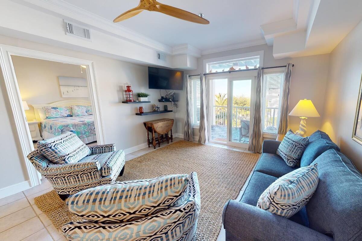 A spacious living room with a large sofa, patterned armchairs, a coffee table, and a TV. There is a balcony with a view, and the room has a modern design with white walls and ceiling.