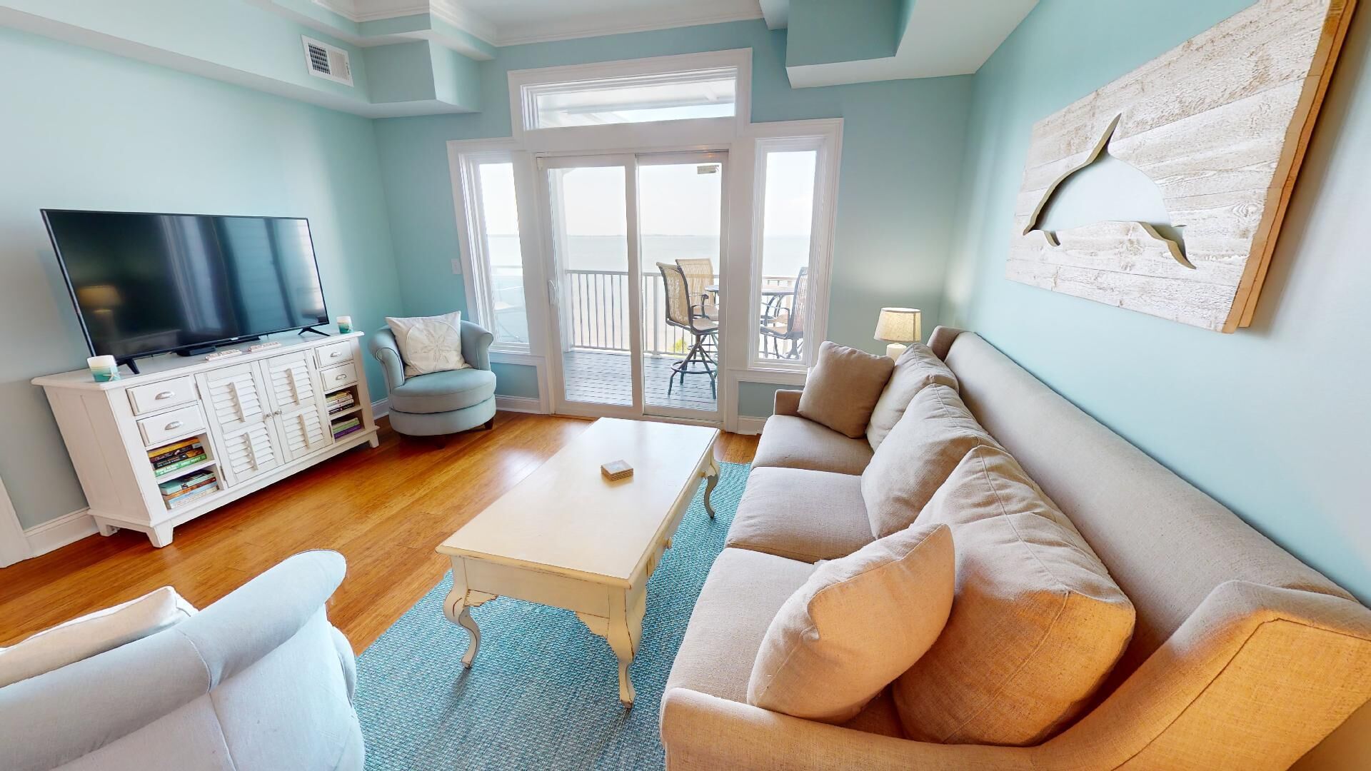 A living room with a television, white furniture, a blue rug, and a view through a glass door to an outdoor area.