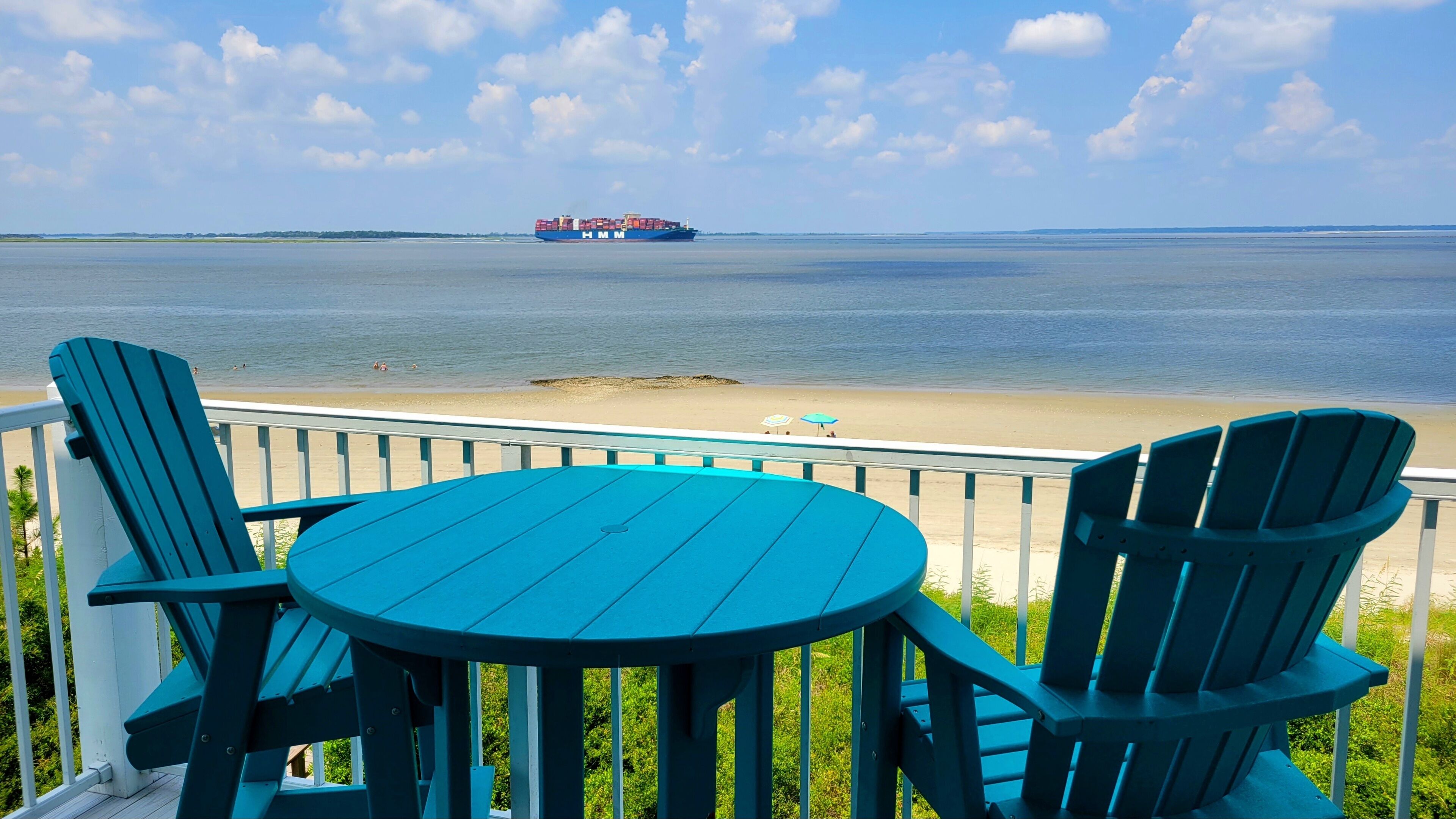 Een terras met een ronde tafel en twee stoelen, met uitzicht op een strand en een vrachtschip op het water.