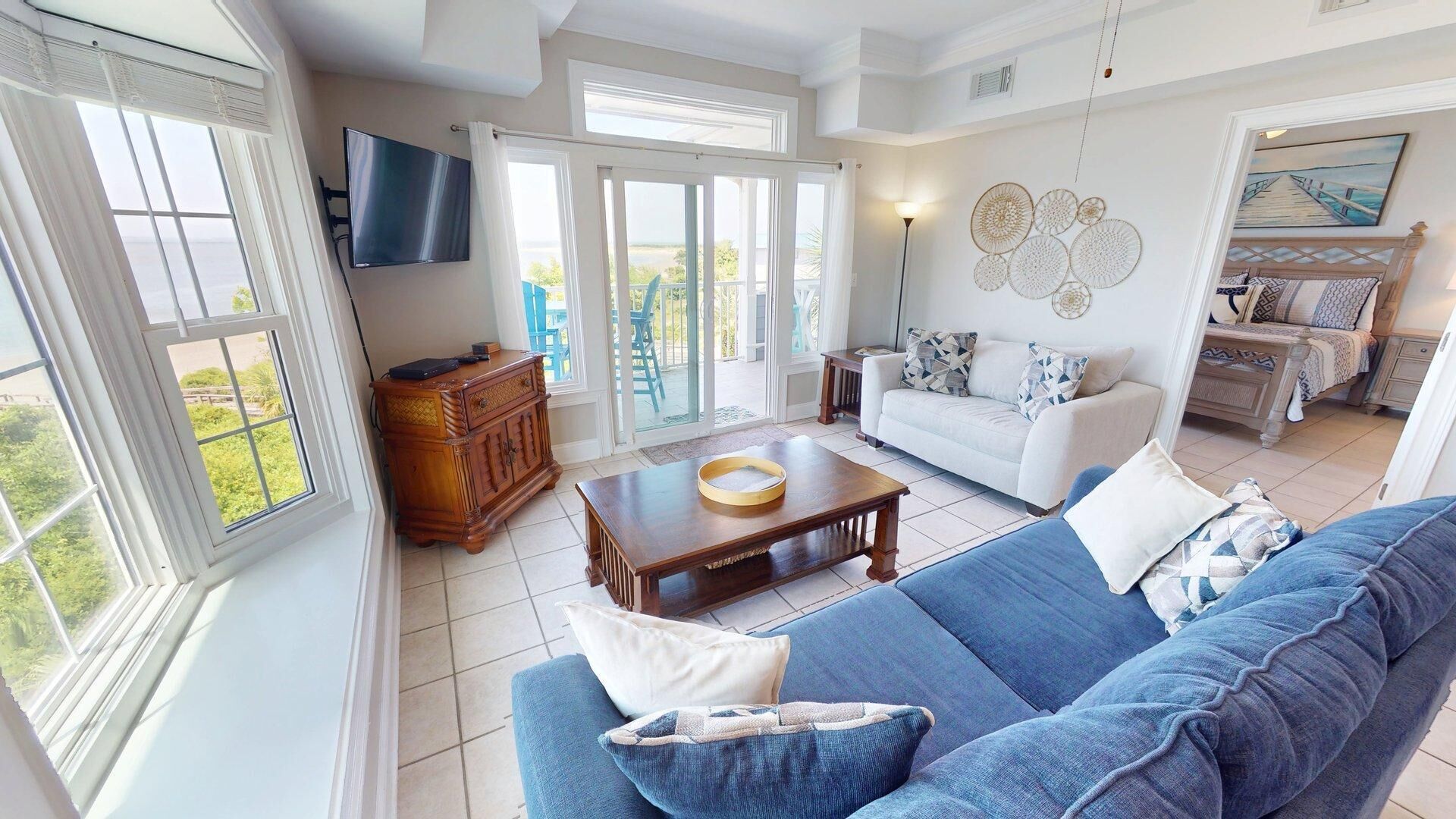 A spacious living area with a blue sofa, a wooden coffee table, and a white couch. There is a television mounted on the wall, and a painting above the bed.