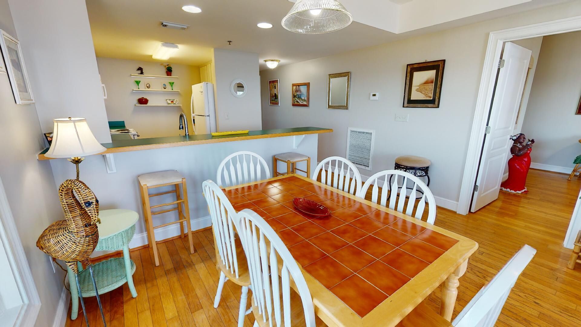 Un coin repas avec une table en bois et des chaises blanches, un bar équipé d’un réfrigérateur, et une cuisine à l’arrière-plan.