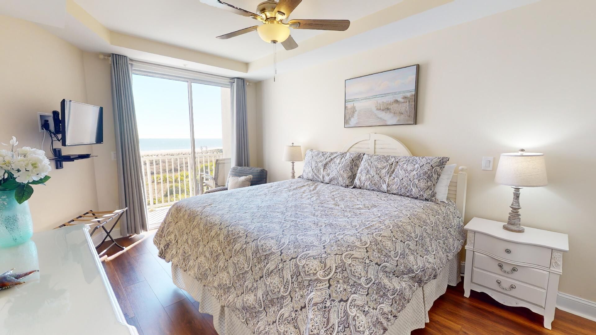 A bedroom with a large bed, a ceiling fan, a painting, a nightstand, a lamp, a window with curtains, and a view of the beach.