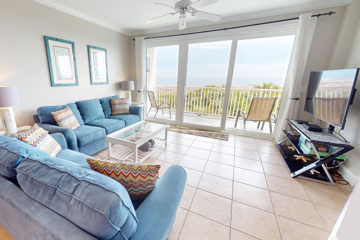 A living room with a blue sofa, a coffee table, and a TV on a stand. There is a balcony with a view of the ocean.
