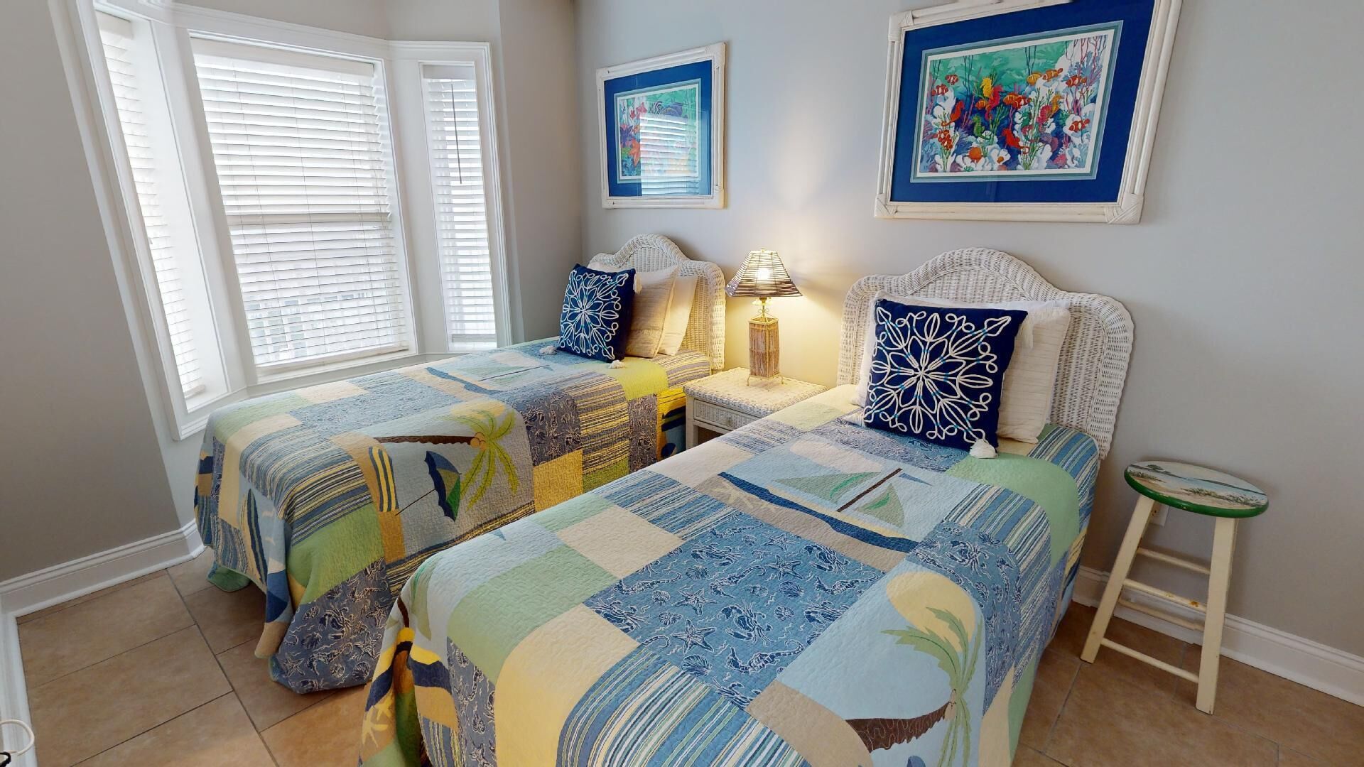 Et soveværelse med to senge, et natbord, en lampe, en lille skammel og to billeder hængende på væggen.