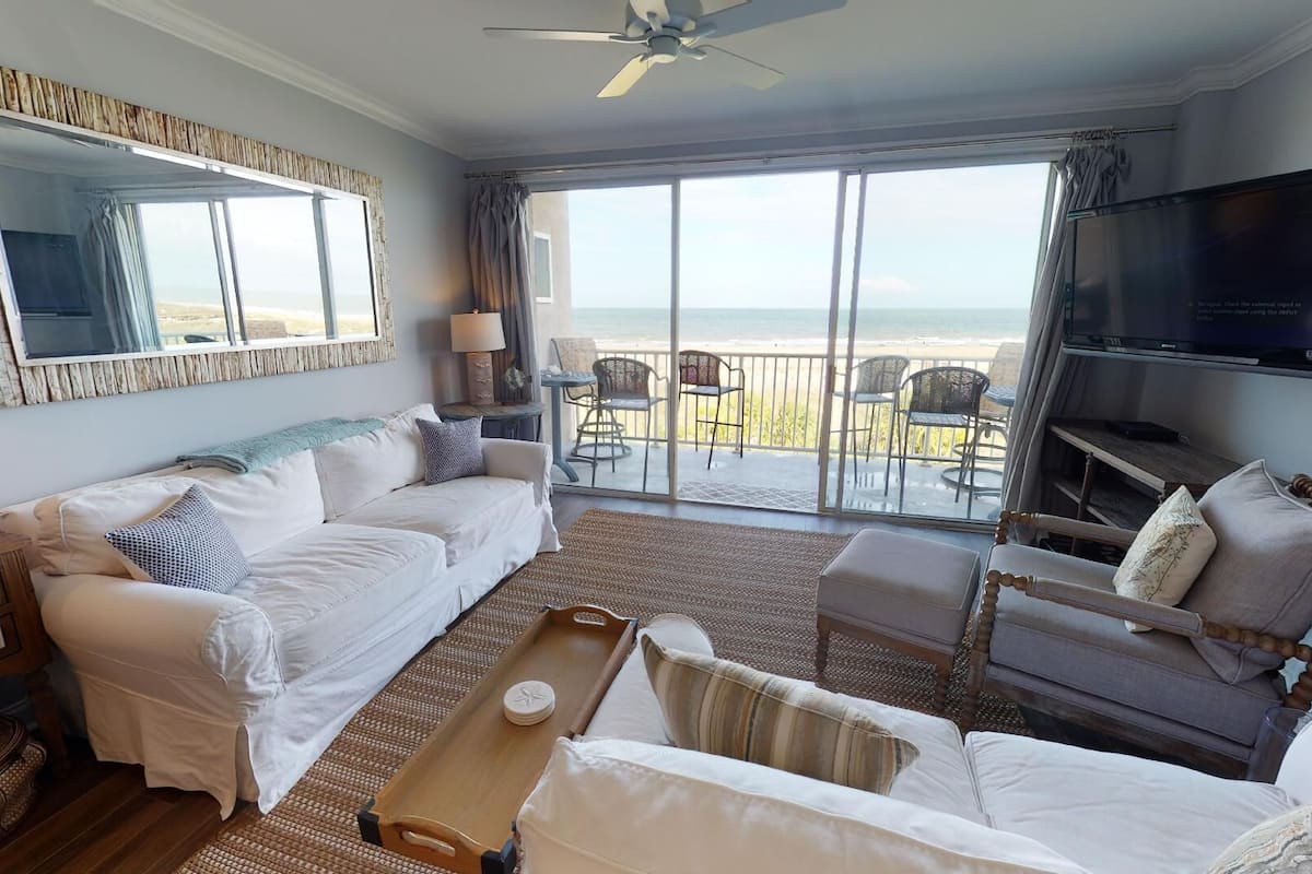 A spacious living room with a large window offering a view of the beach, a ceiling fan, a flat-screen TV, and a cozy seating area with a coffee table.