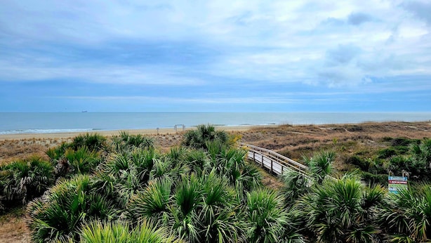 Ein Strand mit einem hölzernen Promenadenweg, einem Schild, das auf ein Zentrum hinweist, und Blick aufs Meer.