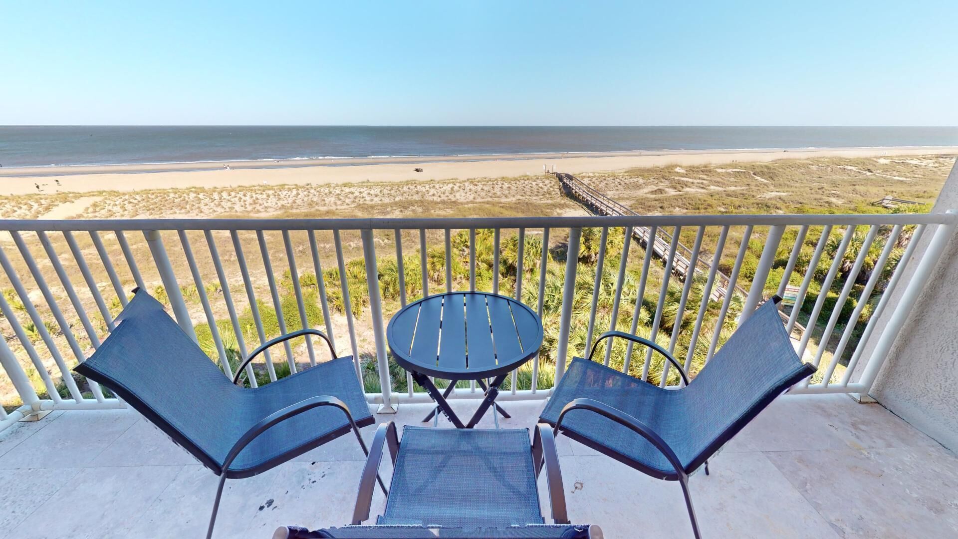 A balcony with two chairs and a small table, overlooking a beach with dunes and a clear sky.