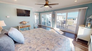A bedroom with a large bed, a ceiling fan, a TV, and a view of the balcony with outdoor furniture. - Beach Ya To It Tybee Vacation Rentals (Tybee Island)