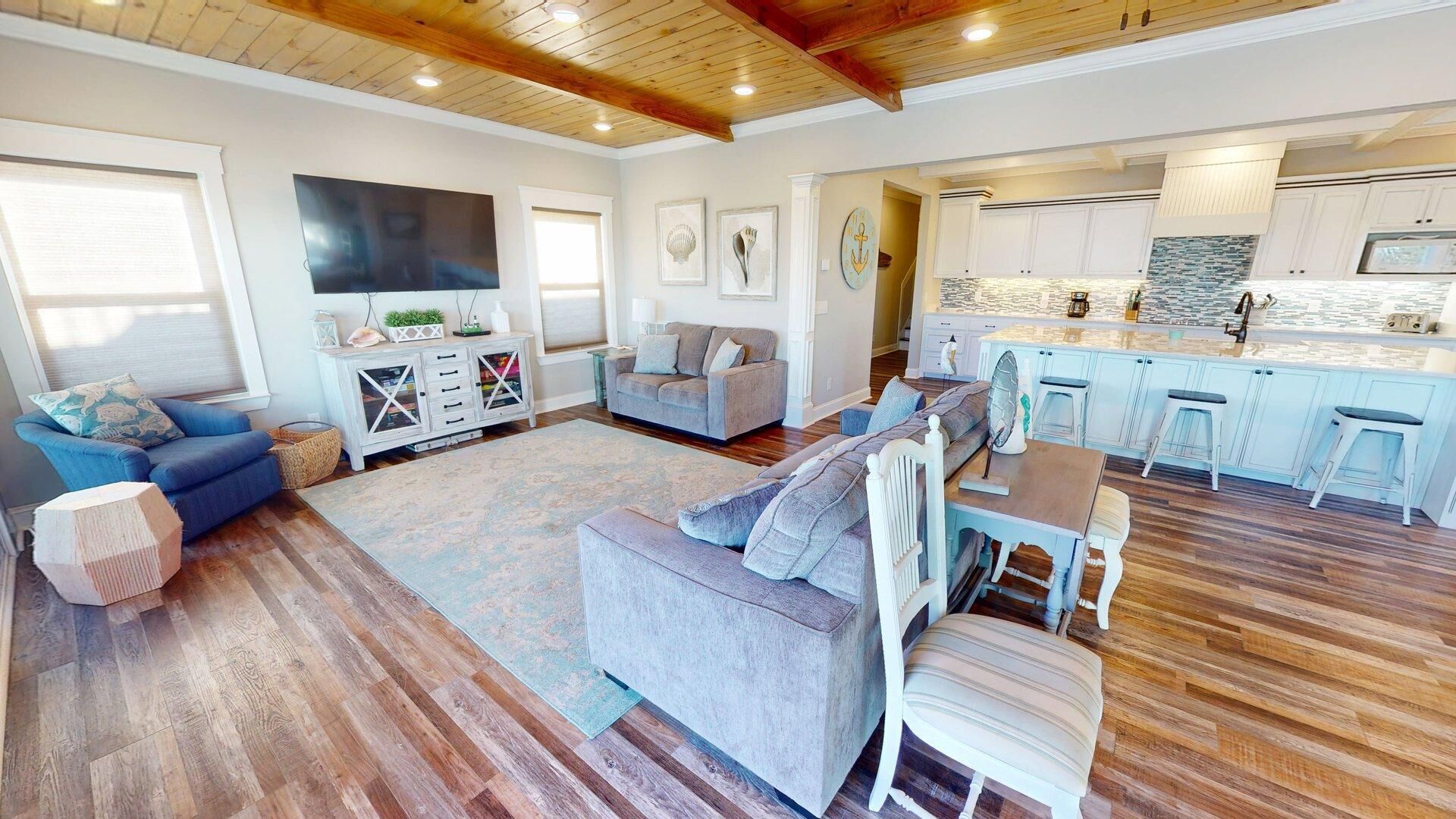 A spacious living room with a grey sofa, a dining table with chairs, and a kitchen area with white cabinets and a stone backsplash.