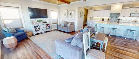 A spacious living room with a grey sofa, a dining table with chairs, and a kitchen area with white cabinets and a stone backsplash.