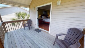 A wooden deck with two Adirondack chairs, a door leading to a bedroom with a bed and two lamps, and a palm tree in the background. - Beachy Keen Tybee Vacation Rentals (Tybee Island)