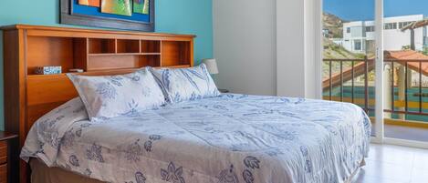 Un dormitorio con cama, mesita de noche, cabecera de madera, un cuadro y un balcón con vista a edificios.