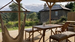 Une terrasse en bois avec un hamac, une table entourée de chaises et, au fond, un bâtiment en pierre.