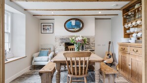 Un coin repas avec une table en bois, des chaises et des bancs. On y trouve une cheminée en pierre surmontée d’un miroir. Une guitare est appuyée contre le mur.