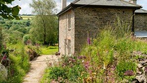 Un bâtiment en pierre, doté d’un toit en pente et de cheminées, entouré d’un jardin où poussent diverses plantes et fleurs.