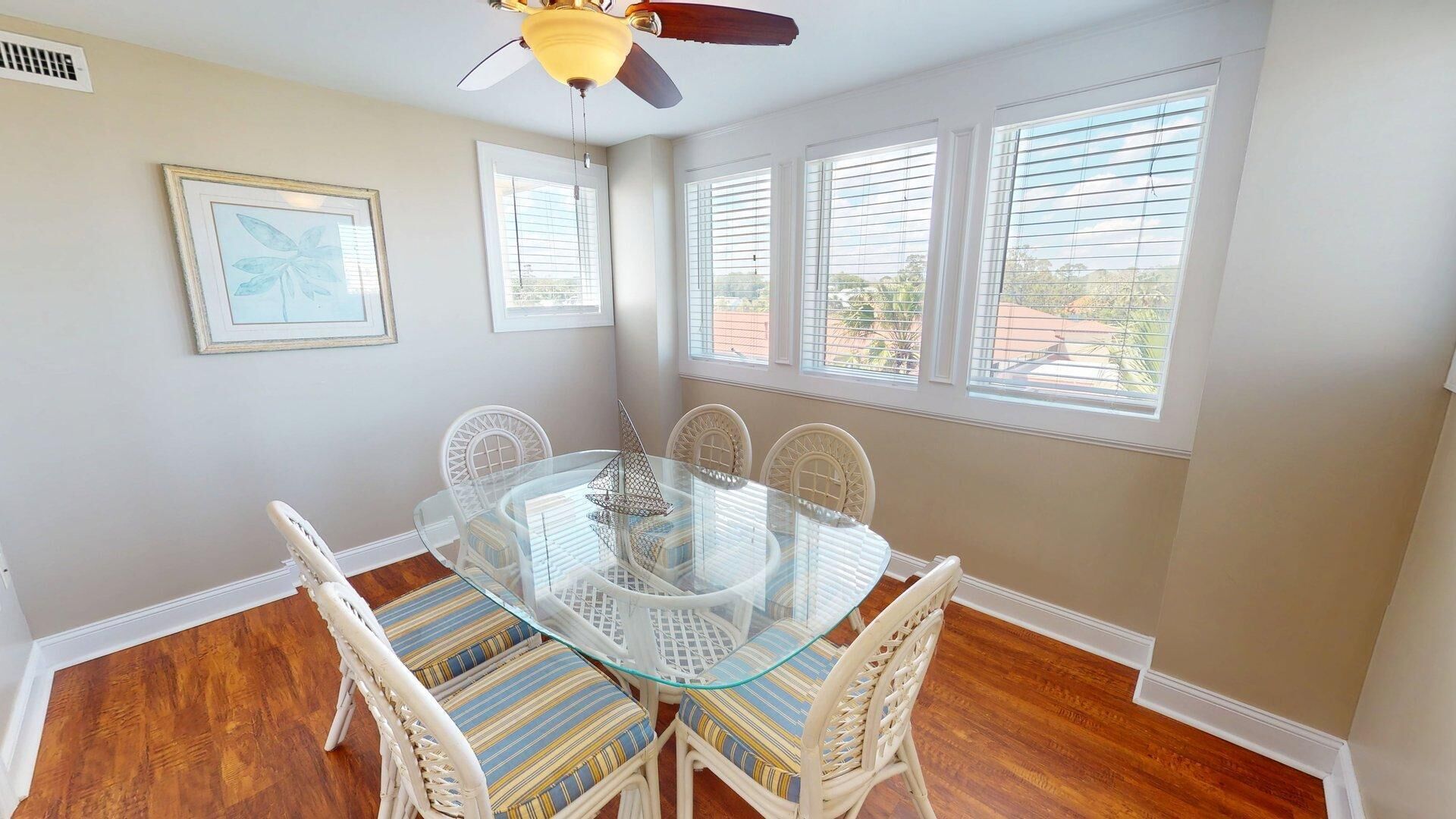 A dining area with a glass-top table surrounded by wicker chairs, white walls, and large windows with blinds.