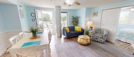 A bright room with a ceiling fan, a blue sofa with yellow cushions, a patterned armchair, a round coffee table, and a dining table with chairs.