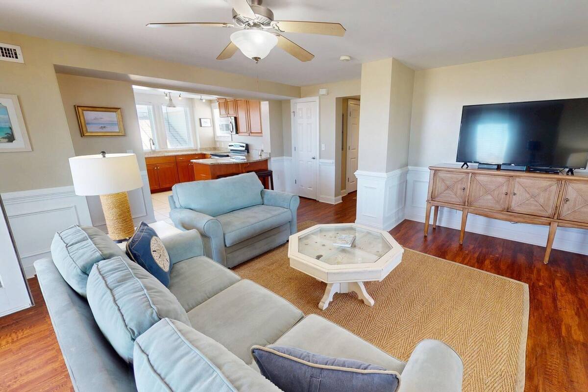 A living room with a sofa, coffee table, and TV stand. There is a kitchen area in the background.