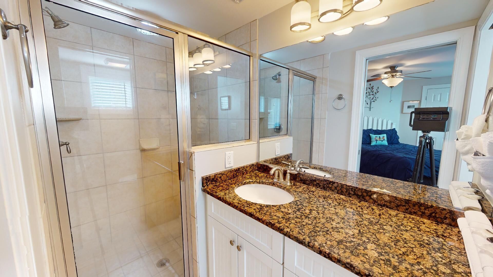A modern bathroom with a glass shower enclosure, a granite countertop, a double sink, and a large mirror.