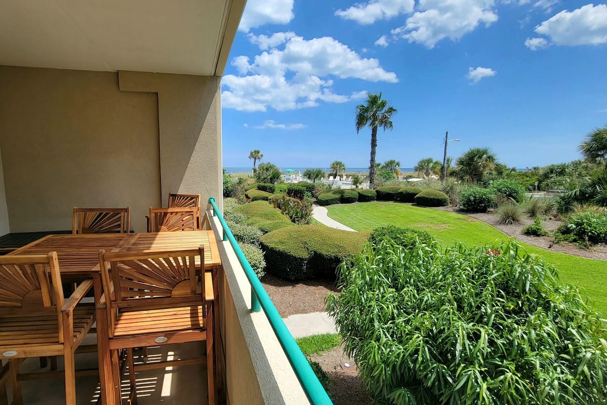 A balcony with a wooden table and chairs, a view of a well-manicured garden with a pathway, and a clear blue sky.